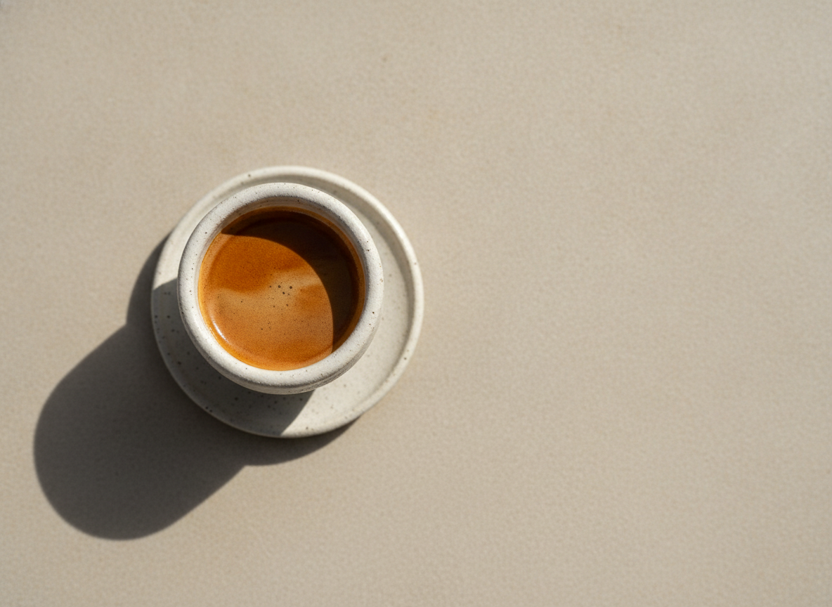 A single espresso in a small ceramic cup on pale stone, shot from above, deep crema catching morning light.